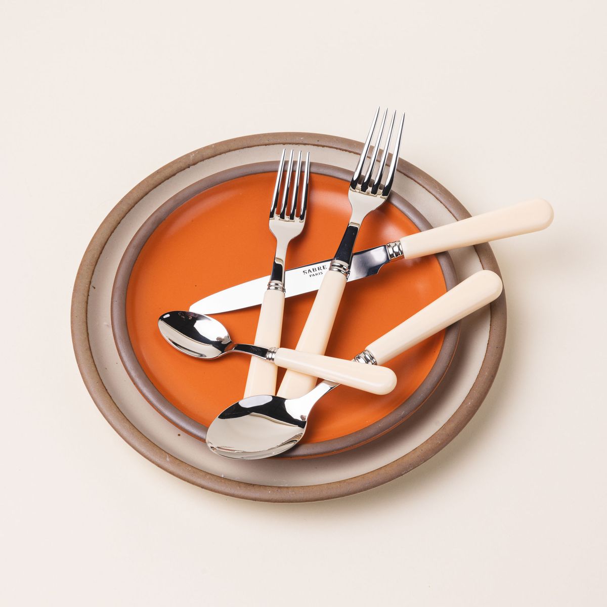 A five piece flatware set with shiny utensils and matte cream handles sits on a bold orange and cream ceramic plates