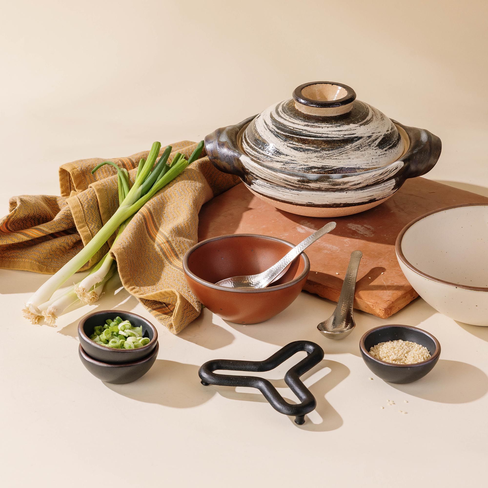 Donabe with bold black-and-white brushwork, styled with serving bowls, utensils, and fresh ingredients on a warm tabletop.
