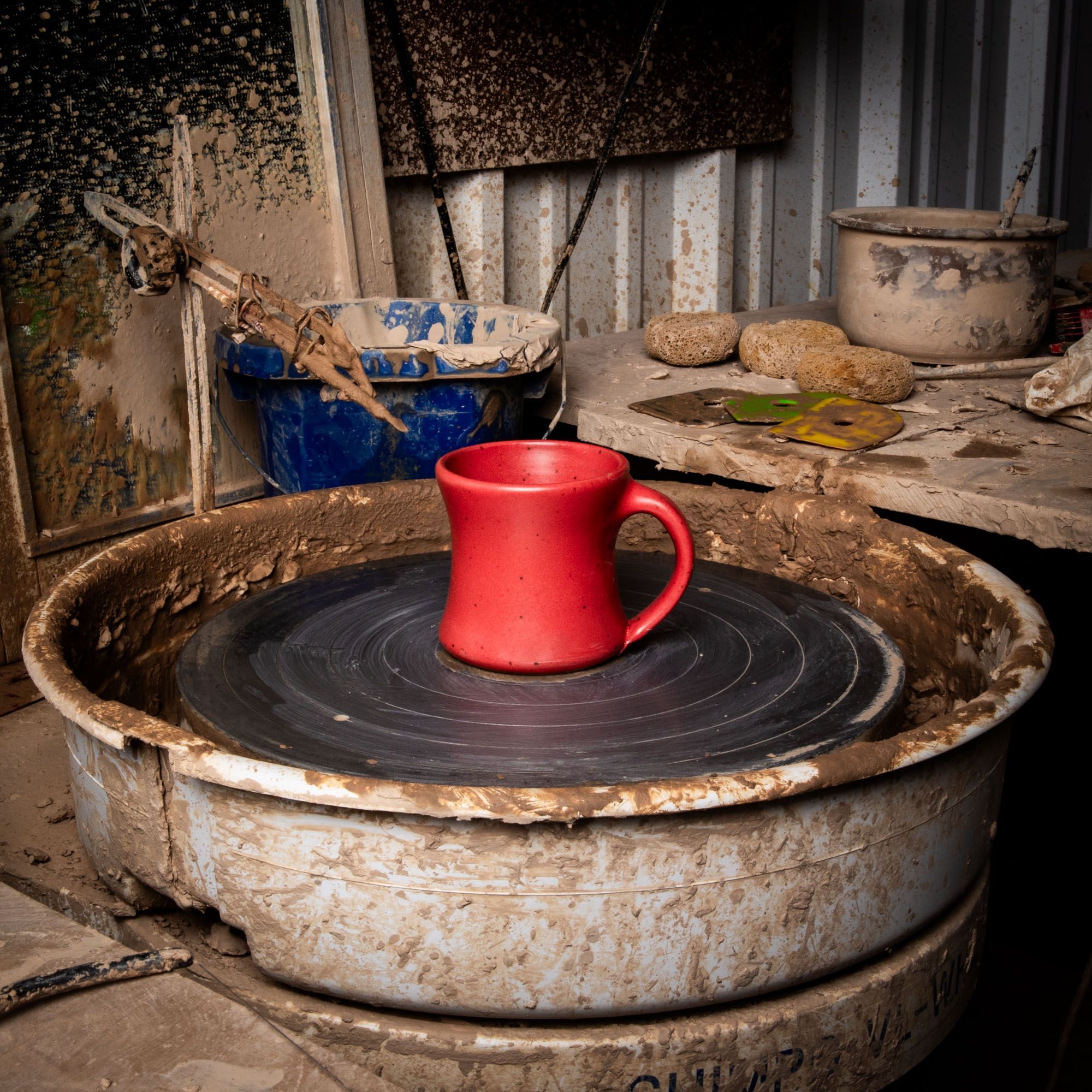 In a pottery studio setting and on a potter's wheel sits a classic diner mug that tapers out on the top and bottom in a bold red color featuring iron speckles and a handle.