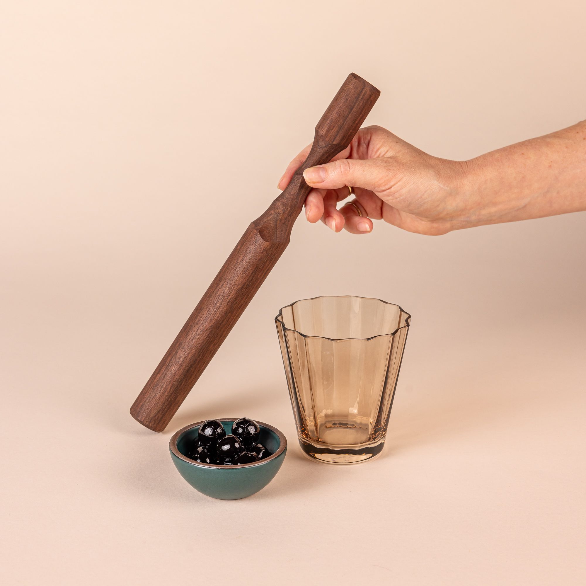 Hand holding a long walnut wood cocktail muddler that tapers slightly towards the handle. Next to it is a short amber ridged lowball glass and a tiny teal ceramic bowl filled with cherries.