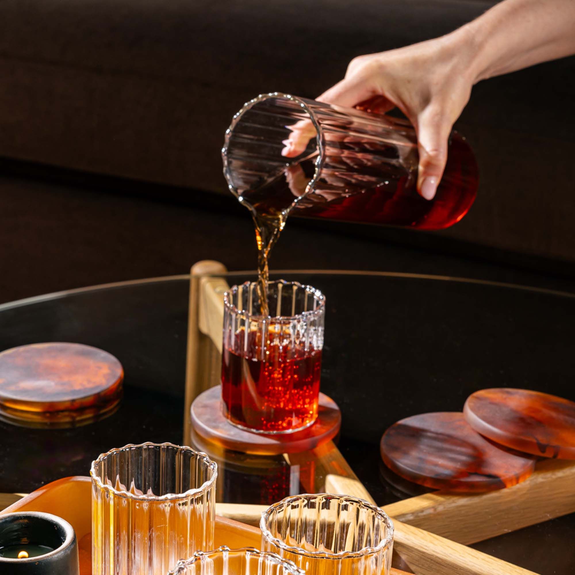 A hand holds a all cylindrical fluted pitcher with a spout on top and is pouring a drink into a short fluted tumbler.