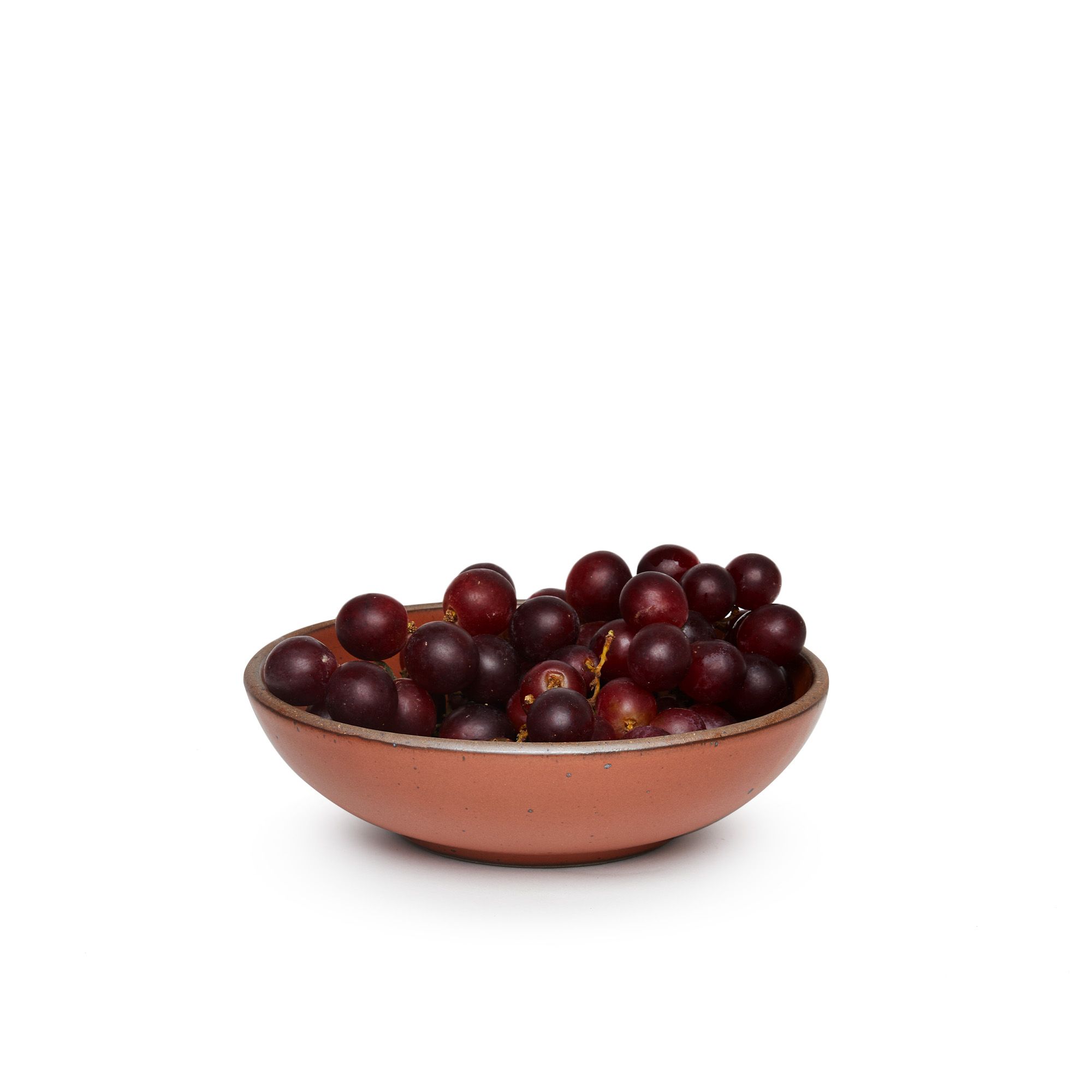 A dinner-sized shallow ceramic bowl in a cool burnt terracotta color featuring iron speckles and an unglazed rim, filled with grapes