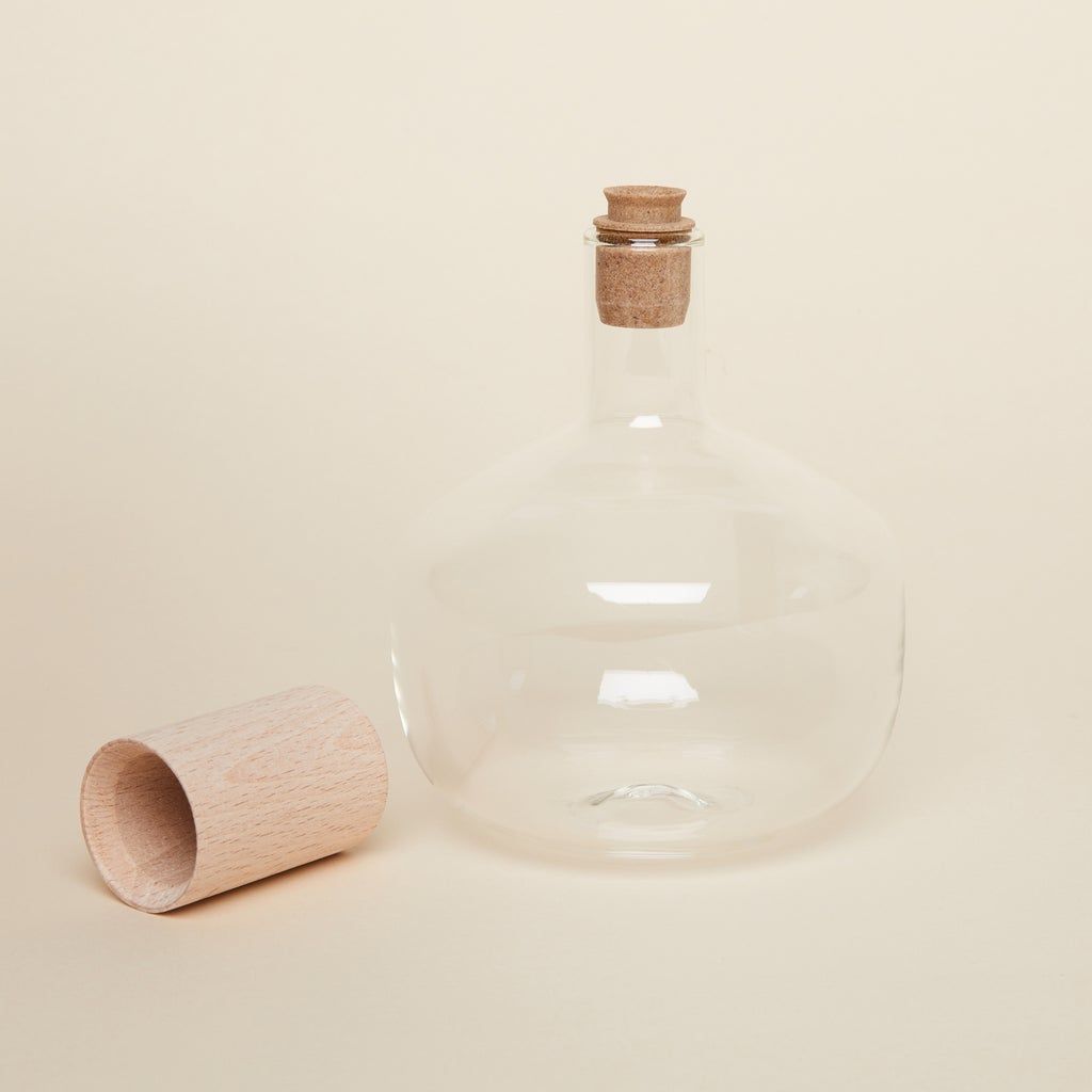 A short glass bottle with wide body with a cork on top and a cylindrical wooden top off to the side