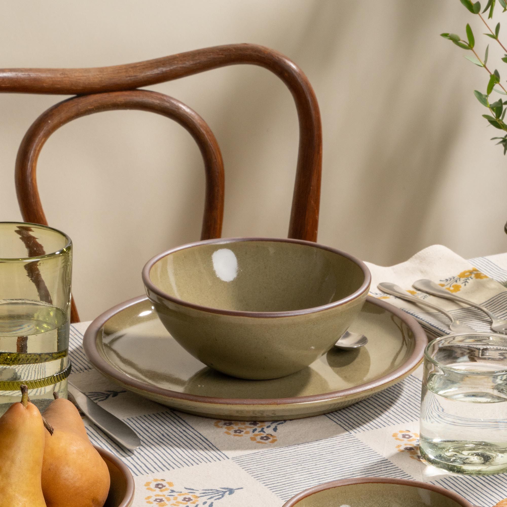 Grey-green stoneware round bowl and plate with water glasses and pears on a striped floral tablecloth.