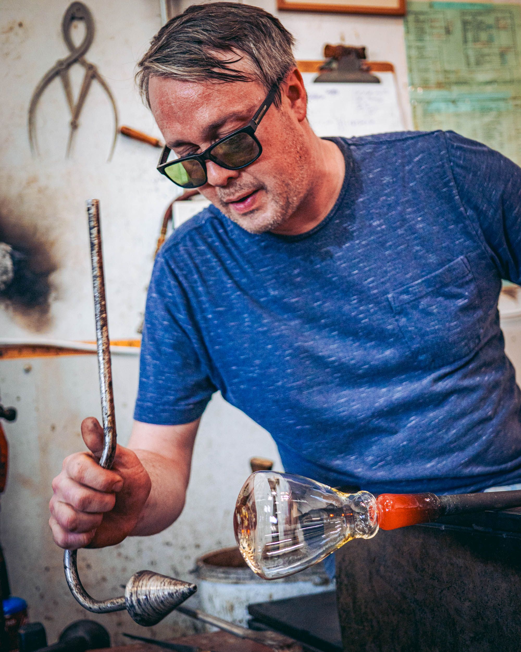 In a studio factory setting, a man is working on blowing glass and is wearing sunglasses.