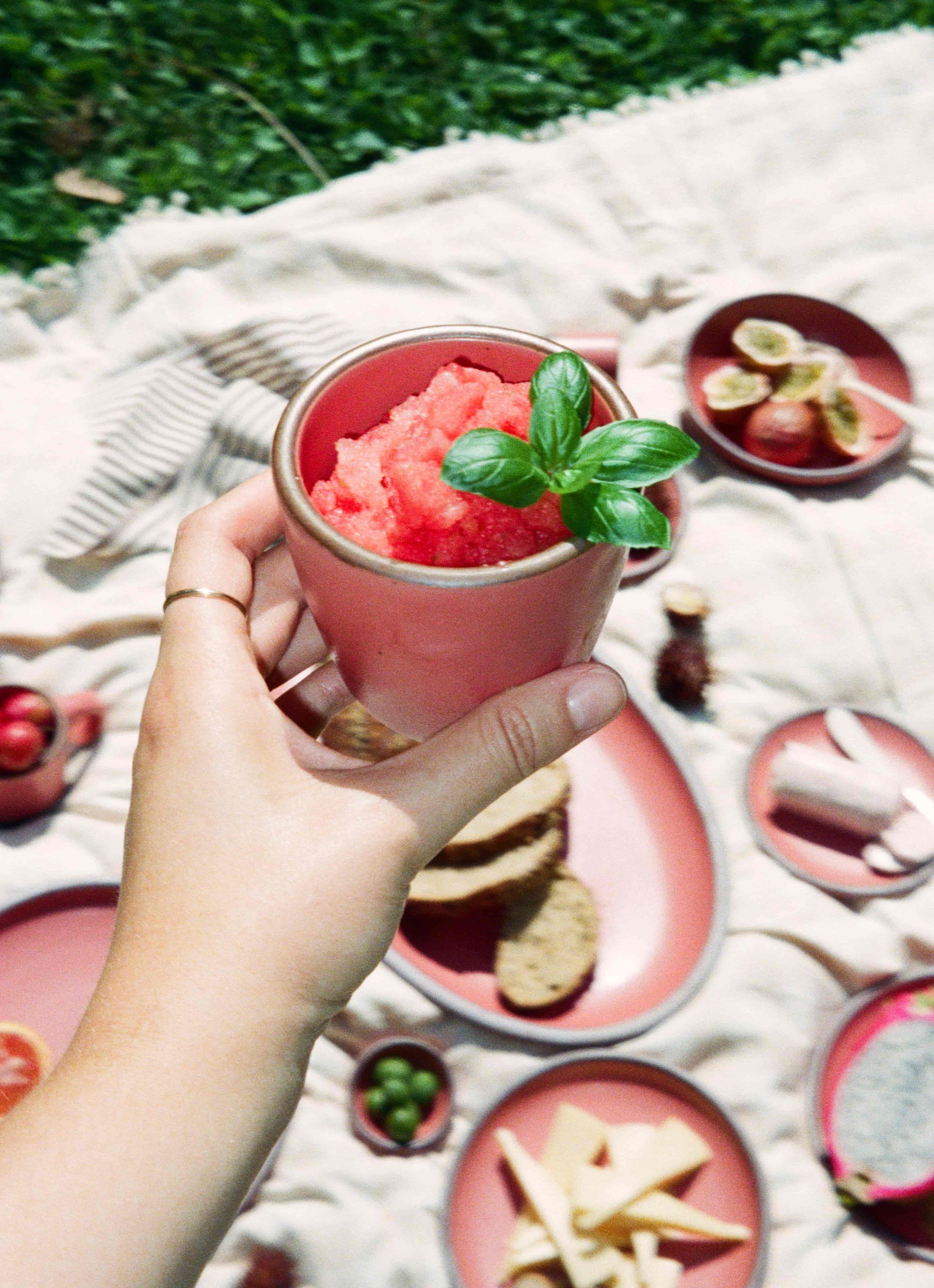 Hand holding a pink ceramic cup filled with bright red granita, topped with fresh basil, over a picnic spread on a cream blanket.