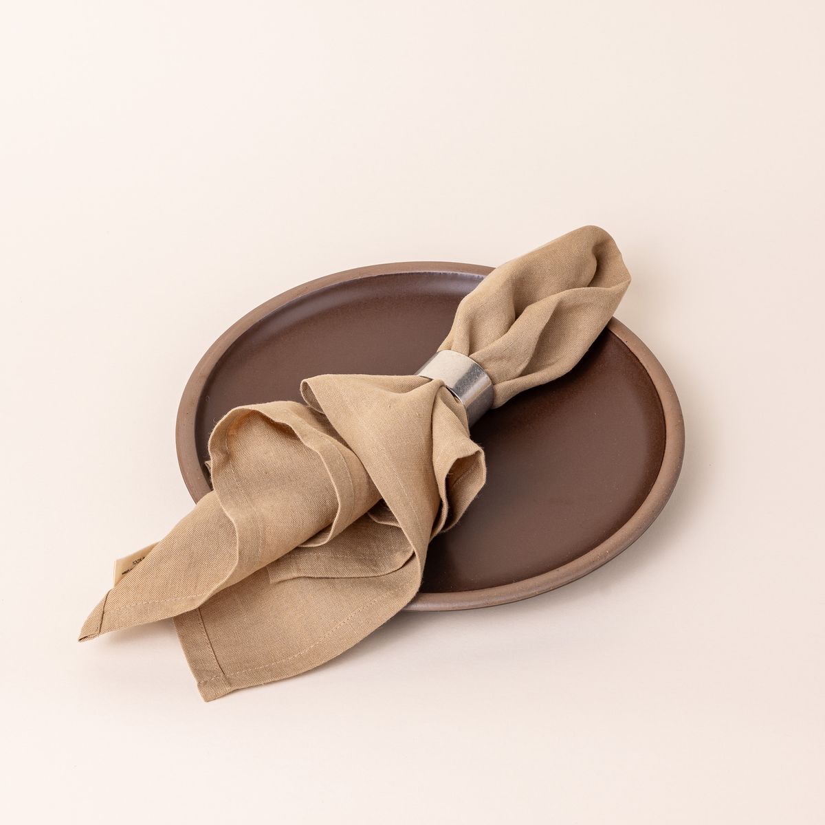 A light tan linen napkin is folded by the center through simple pewter napkin ring and sitting on a brown dinner plate.