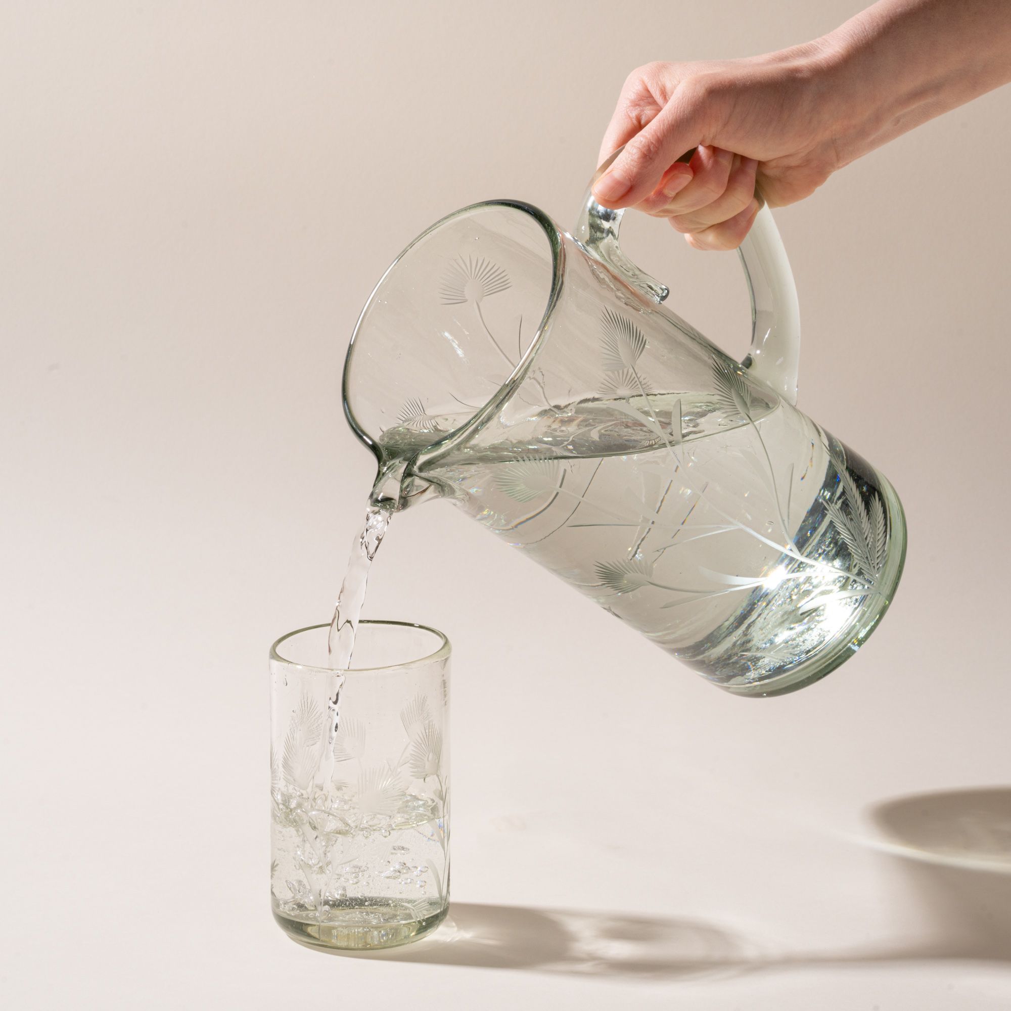 A hand holds a pitcher and pours water in a tumbler, both pieces are clear, made of recycled glass, and have etched thistle designs on them.