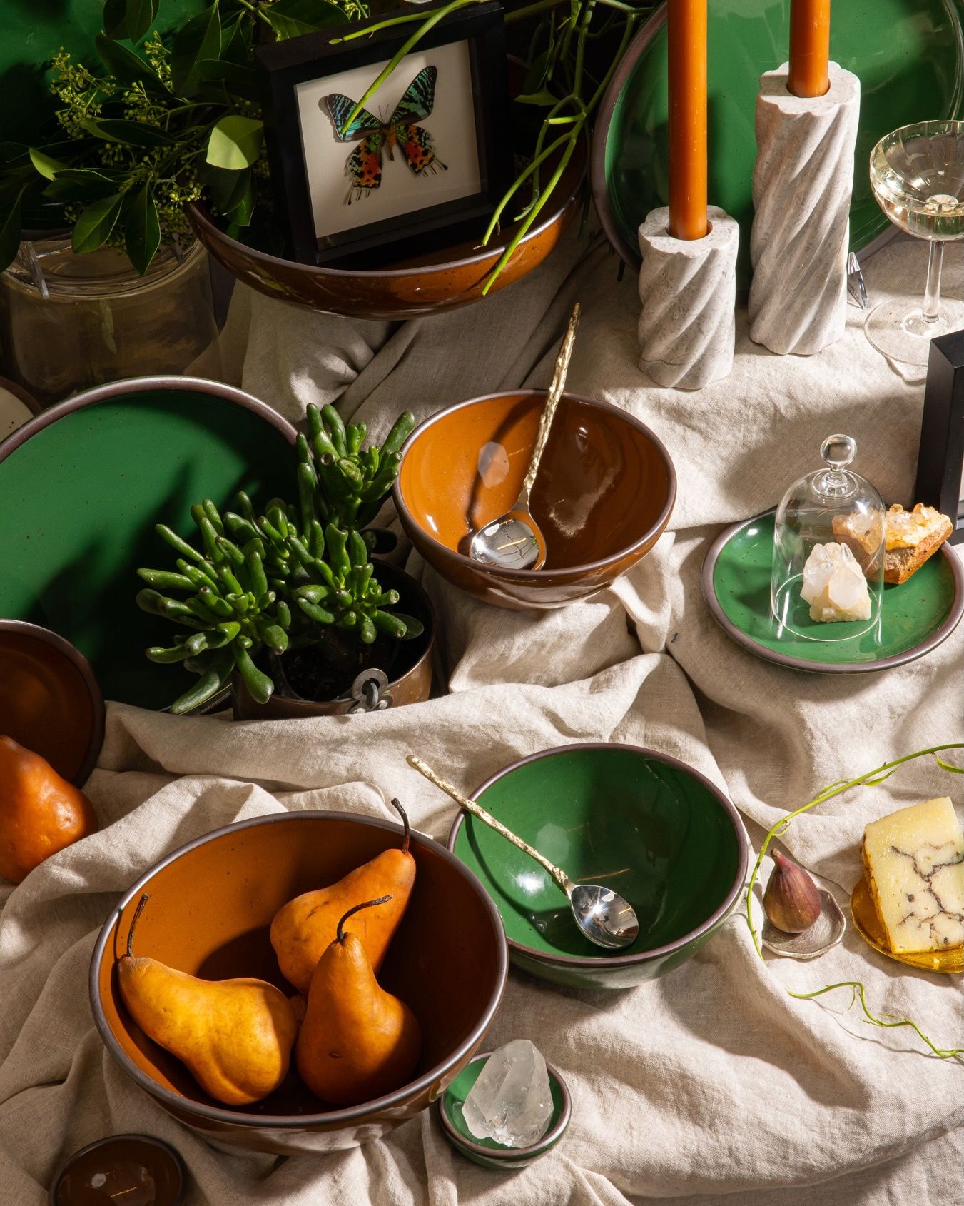 Glossy green and brown ceramic bowls with pears, cheese, crystals, candles, and framed butterfly on a draped linen table.