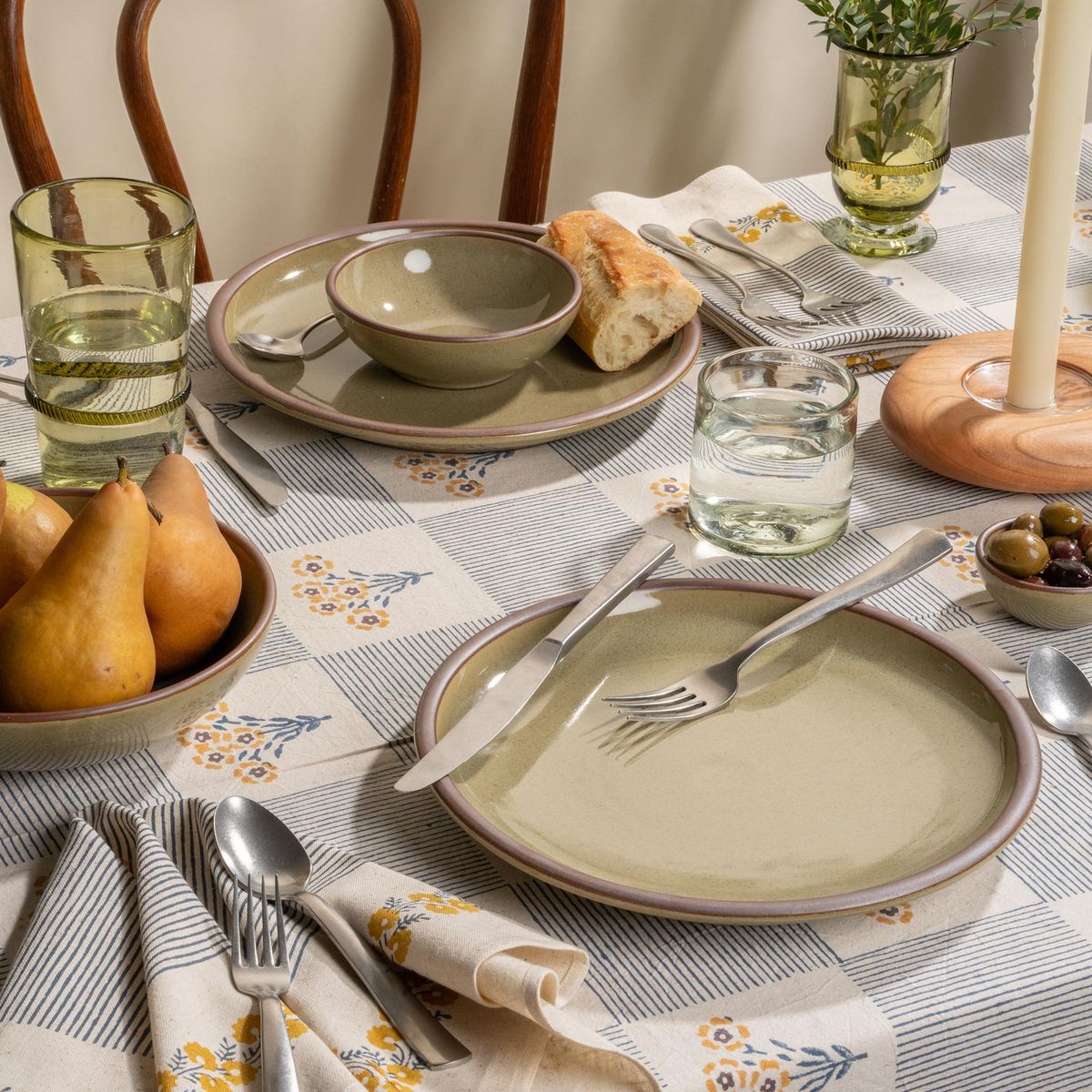 Table filled with grey-green dinnerware with pears, bread, olives, and water on a striped floral tablecloth.