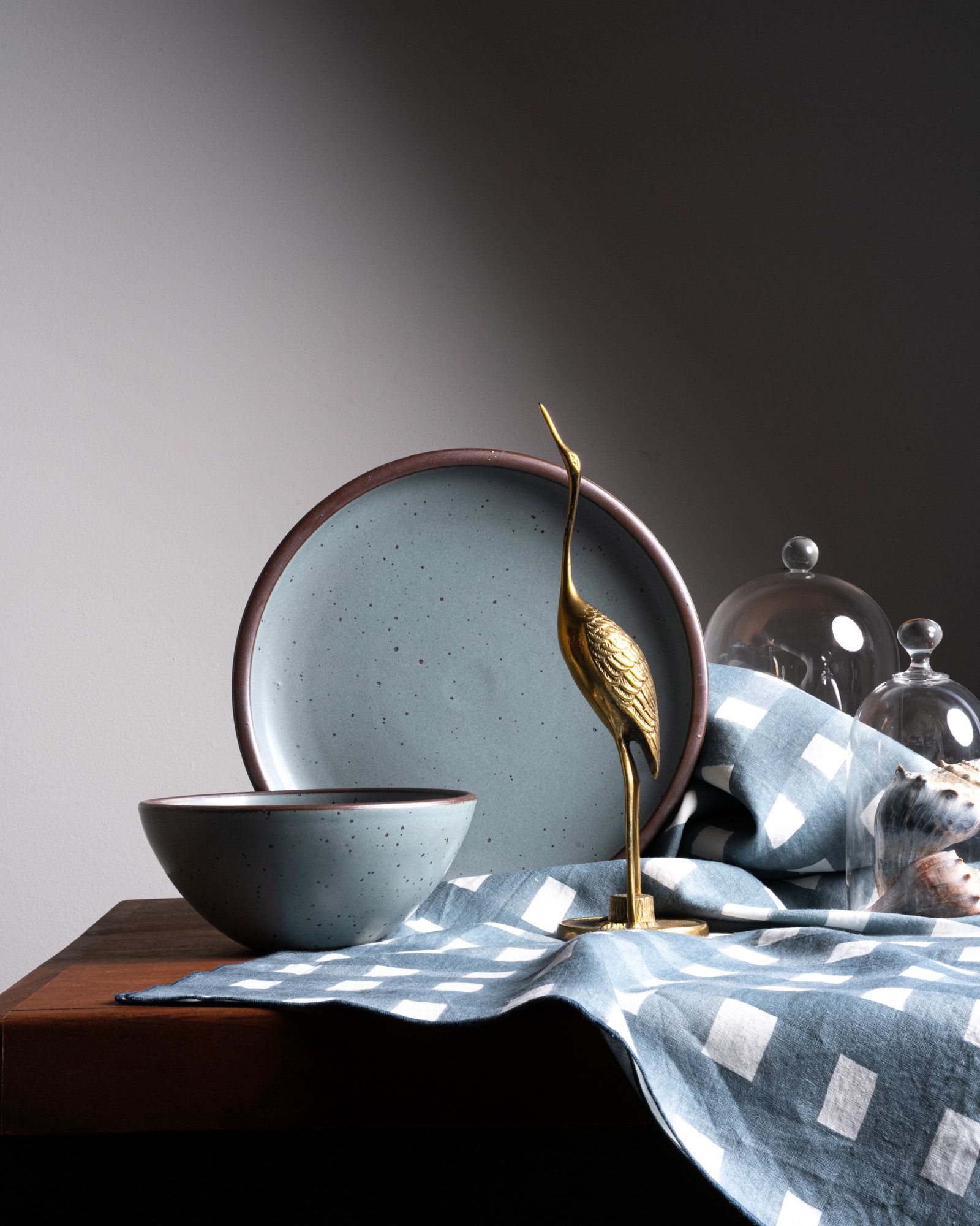A round ceramic bowl and propped dinner plate in a blue-grey color, with an artfully arranged brass bird and crosshatch tablecloth