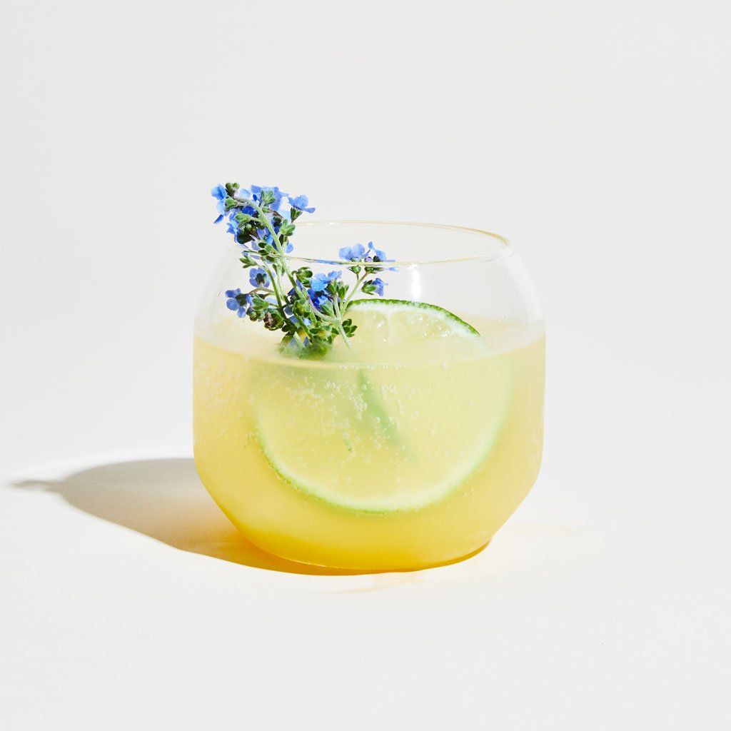 A stemless wine glass with a rounded shape, containing yellow liquid, a slice of lime and a spring of blue flowers