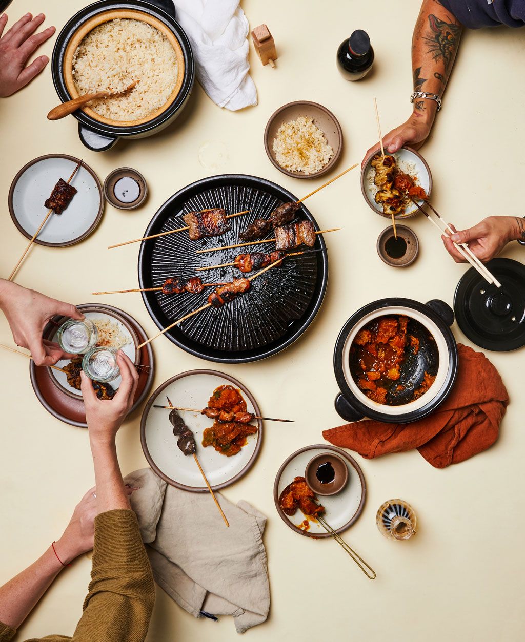 An overhead view of people partaking in a feast with kebabs on a cast iron plate, a donabe with rice, and other foods with white plates and dinner napkins.