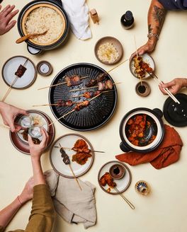 An overhead view of people partaking in a feast with kebabs on a cast iron plate, a donabe with rice, and other foods with white plates and dinner napkins.