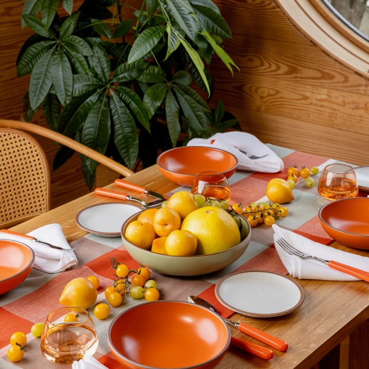 A table set with a gingham table runner, bold orange bowls and cool white plates, orange-handled flatware, whiskey snifters, and a centerpiece of fruits and vegetables. Also pictured is a woven chair behind the table and a large houseplant in the background