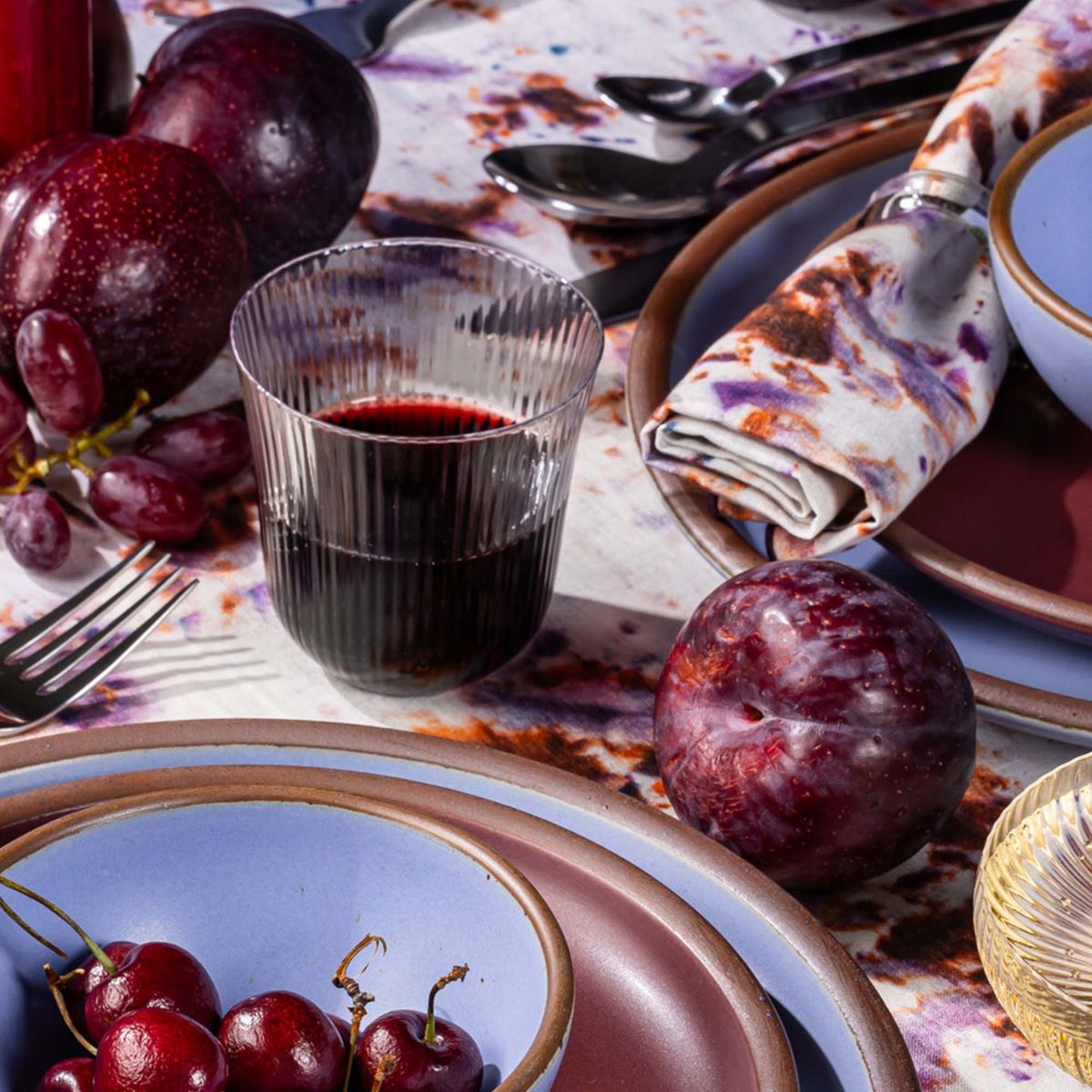 A ribbed glass that curves out on top filled with red wine and sitting next to place settings with periwinkle ceramic plates