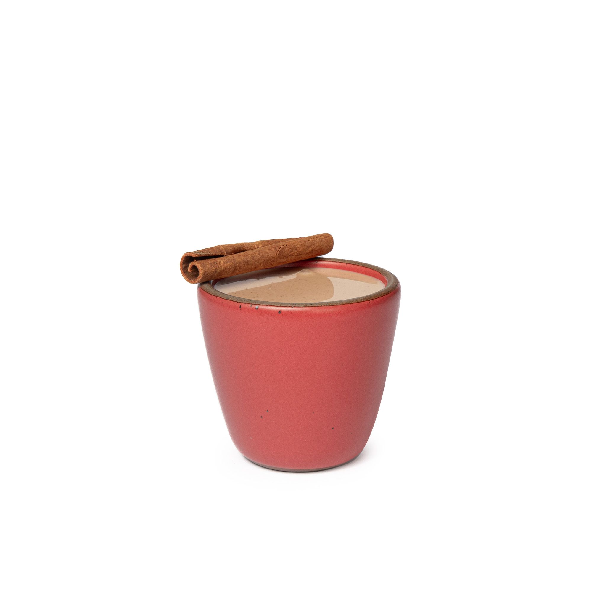 A short cup that tapers out to get wider at the top in a bold red color featuring iron speckles. The cup is filled with a buttered chai drink with a cinnamon stick on top. 