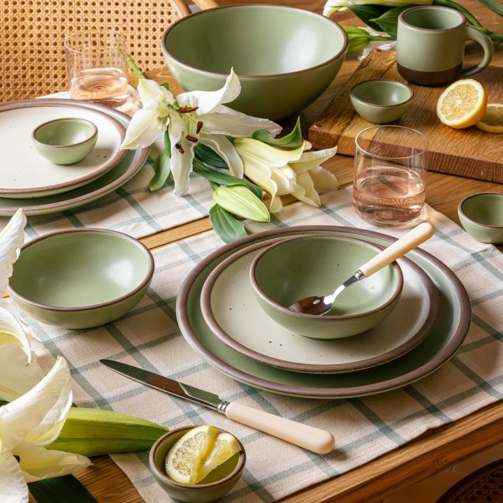 A table set with a gingham rectangular placemats in cream, off-white, and sage green colors, sage green plate, cream handled flatware, glasses, and lilies.