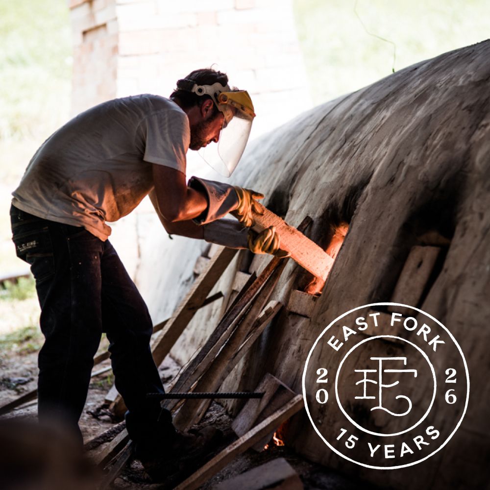 Alex putting wood into the window of an outdoor wood-fired kiln. An overlay stamp is on the corner of the photo that reads "East Fork 2026 15 Years".