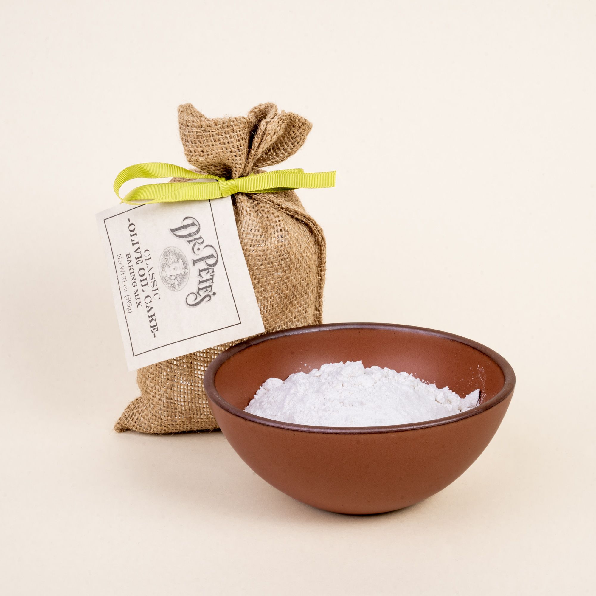 A burlap bag with a lime green ribbon and a label that reads "Dr. Pete's Classic Olive Oil Cake Mix' with a large bowl filled with baking mix.