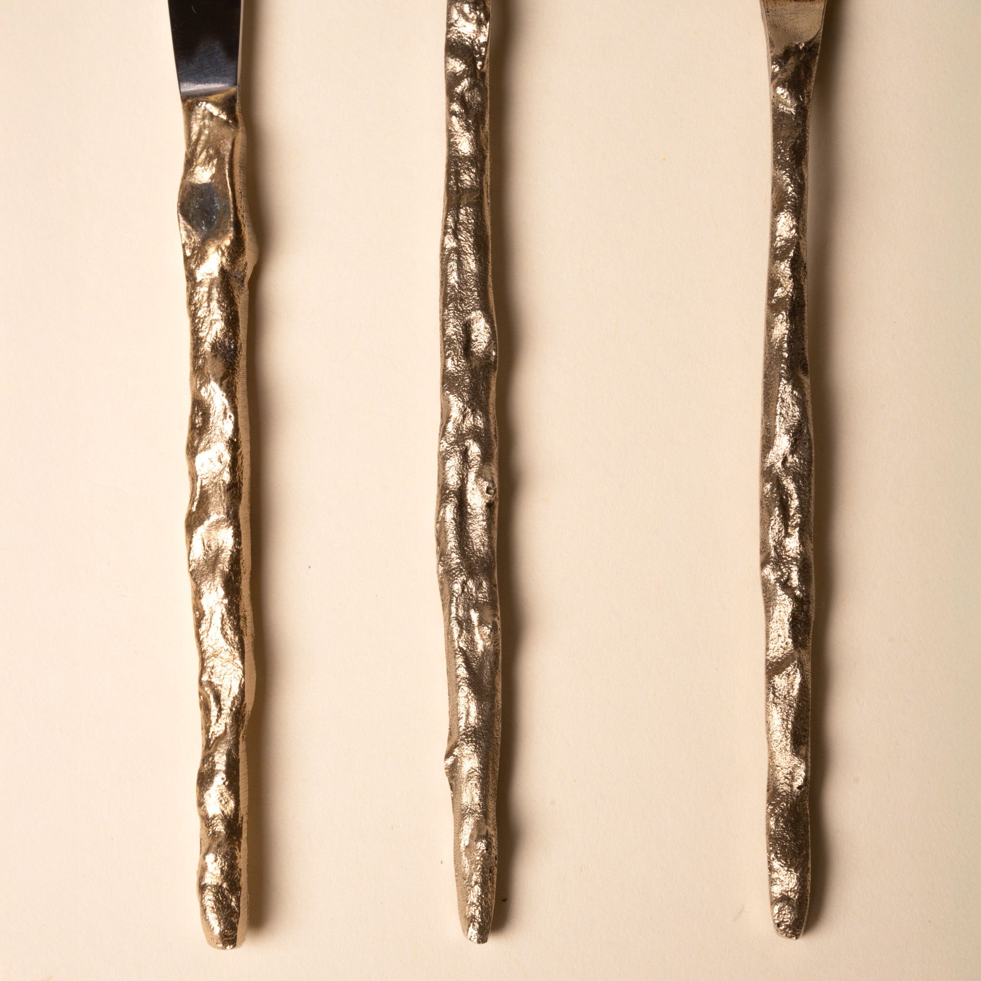Close-up of three hammered metal utensil handles with textured, organic shapes on a beige background.