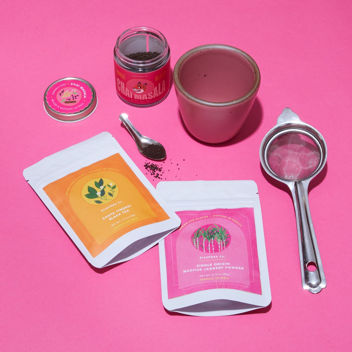 Two Chai Kuhlads – handleless mugs – in Rococo, a hot pink seasonal glaze made by East Fork Pottery. Photographed with two packages of chai mix. Photographed with a stainless tea strainer, a small glass container with a hot pink label holding Diaspora Co.'s house chai blend and two white plastic bags of more tea from Diaspora. Each have a bright label, in orange and pink.