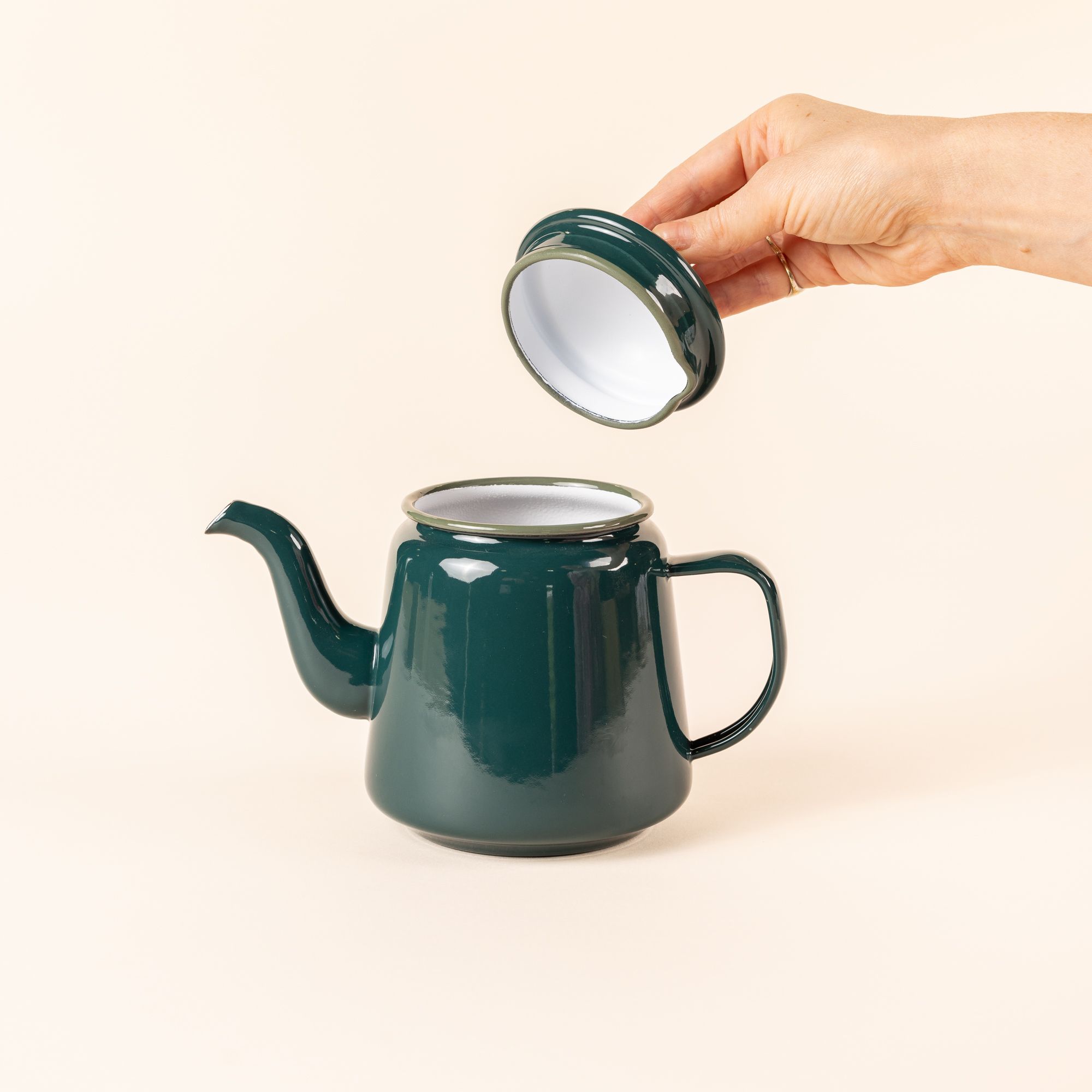 A hand holds the lid above an open enamel teapot. The teapot is deep dark teal enamel with a thin handle, large spout, and simple lid