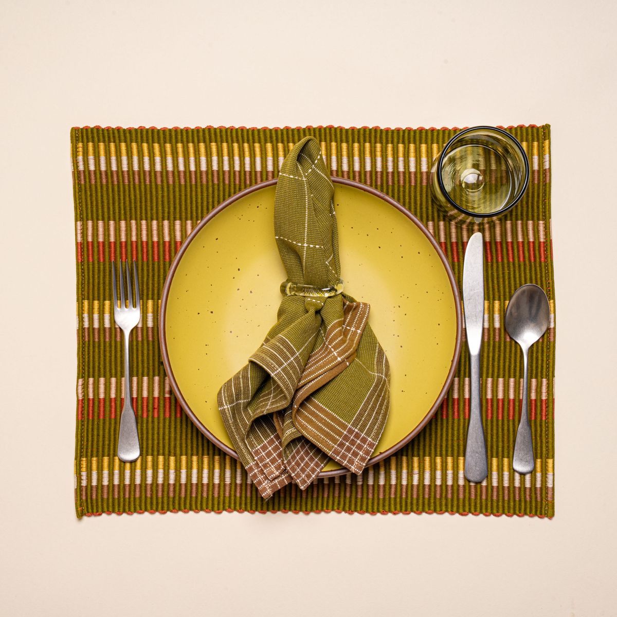 A yellow ceramic plate with silverware, glass, a green grid napkin on a rectangle placemat with offset stripe design that is hand woven in olive, tan, orange, and yellow colors