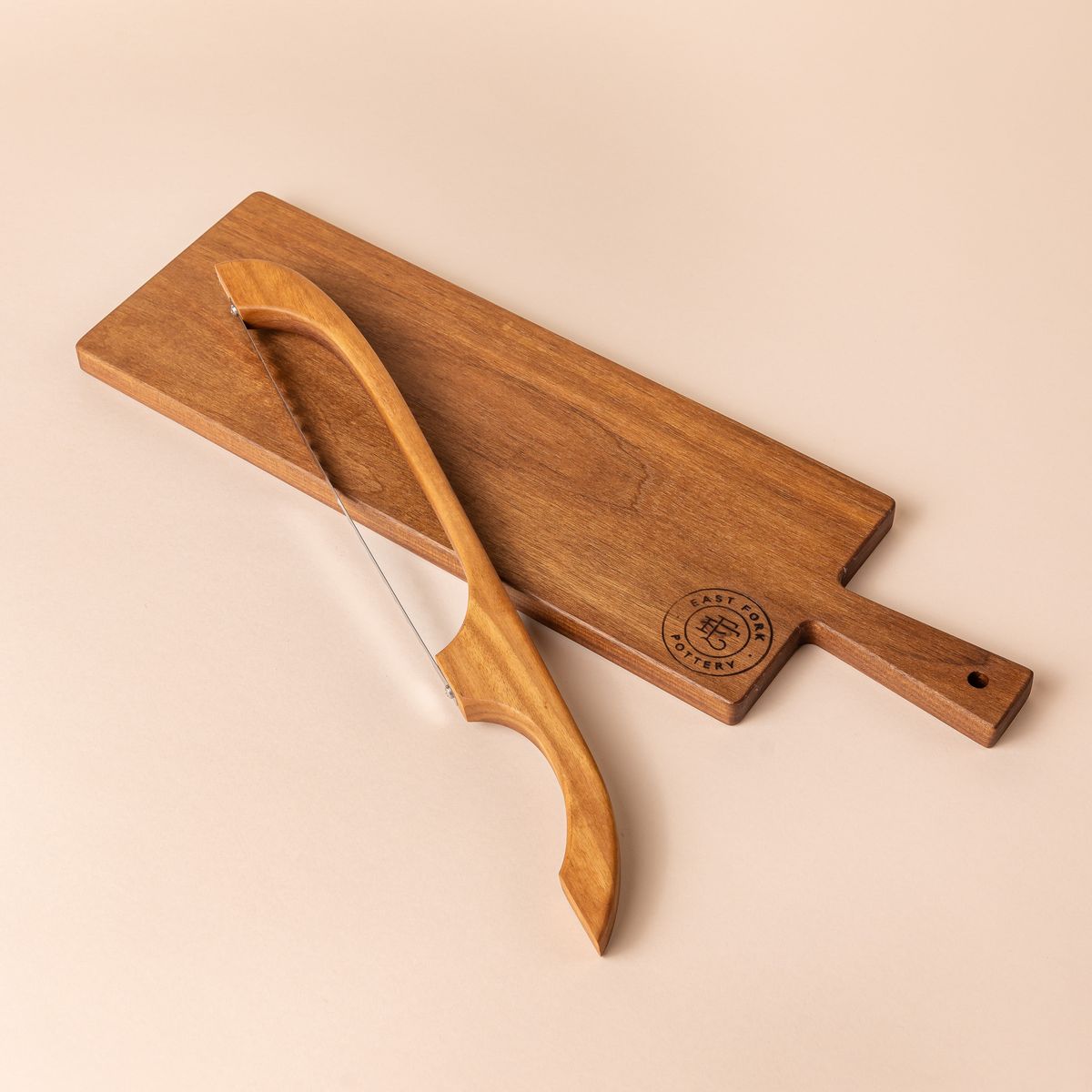 A wood rectangle bread board with an "East Fork Pottery" stamp on the corner and a handle. A matching wood bread bow sits on top of it.