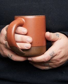A person wearing a black sweatshirt is holding a ceramic terracotta mug with 2 hands.