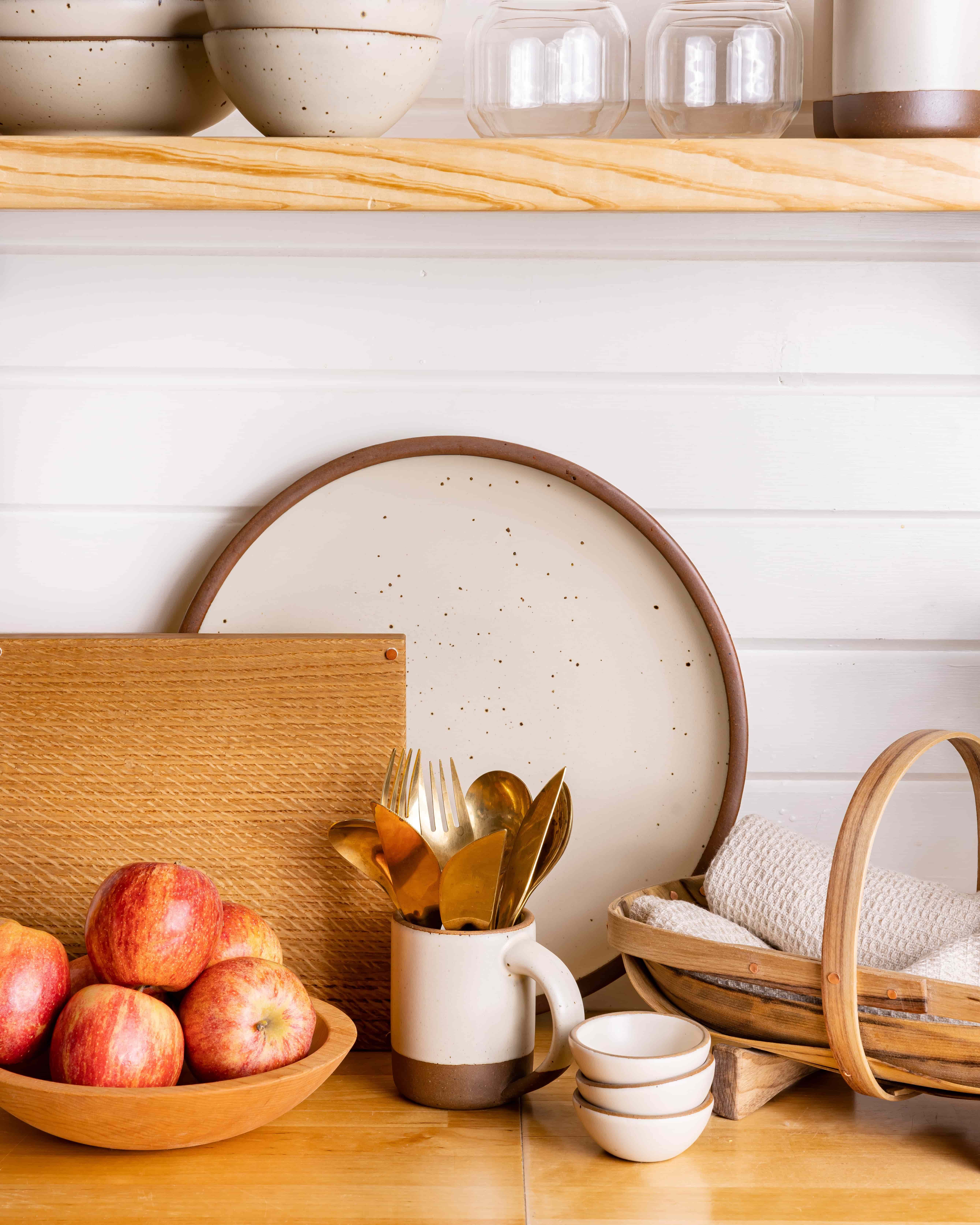 On a kitchen counter is a wooden trug with rolled up napkins in it, a large serving platter, a cutting board, and bowl filled with apples.