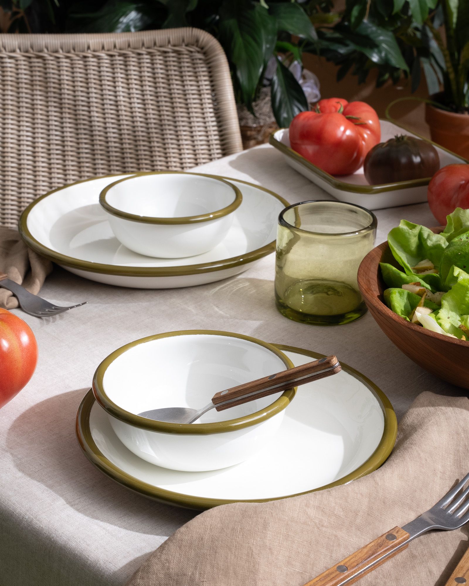 Enamel plates and bowls on a table in a white color with an olive green rim, alongside green glassware, and silverware with wood handles.