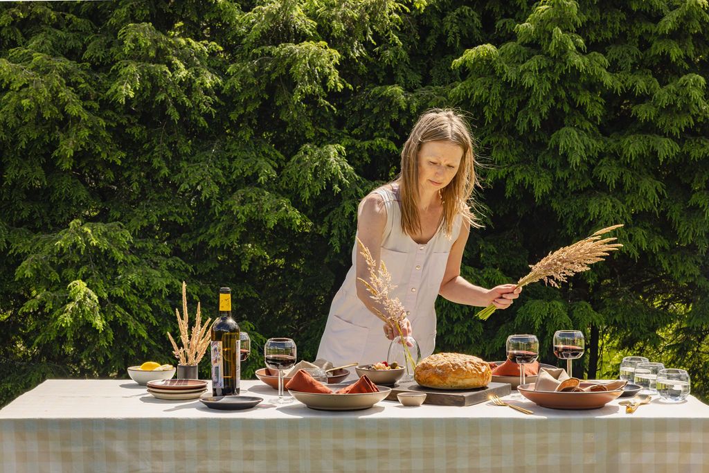 At an outside table, someone is arranging dried florals for a centerpiece. The table is filled with plates, wine glasses, a bottle of wine, loaf of bread, and more.