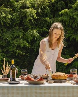 At an outside table, someone is arranging dried florals for a centerpiece. The table is filled with plates, wine glasses, a bottle of wine, loaf of bread, and more.