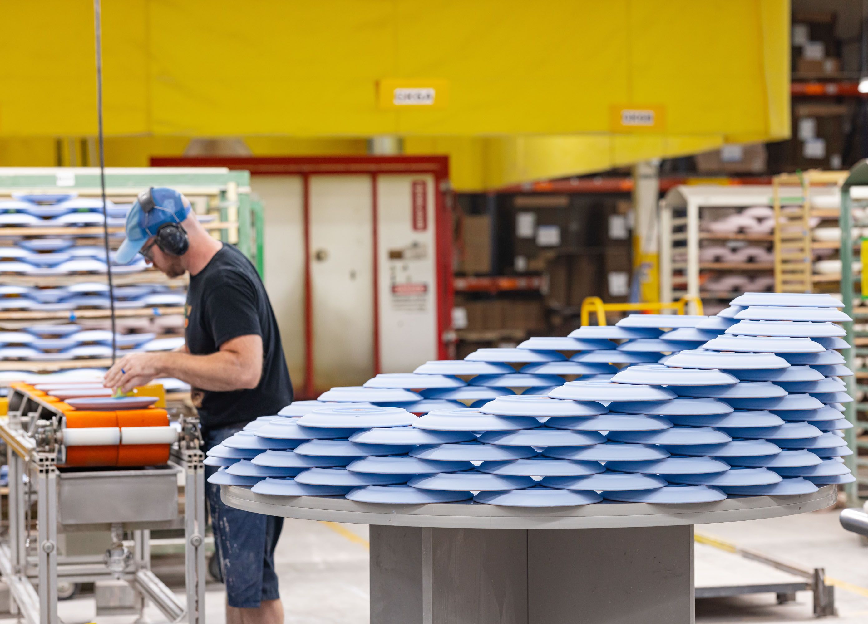In the foreground is a masterful honeycomb stack of freshly glazed ceramic plates drying, in a bright light periwinkle shade. In the background a man works on finishing touches for the next glazed plate.