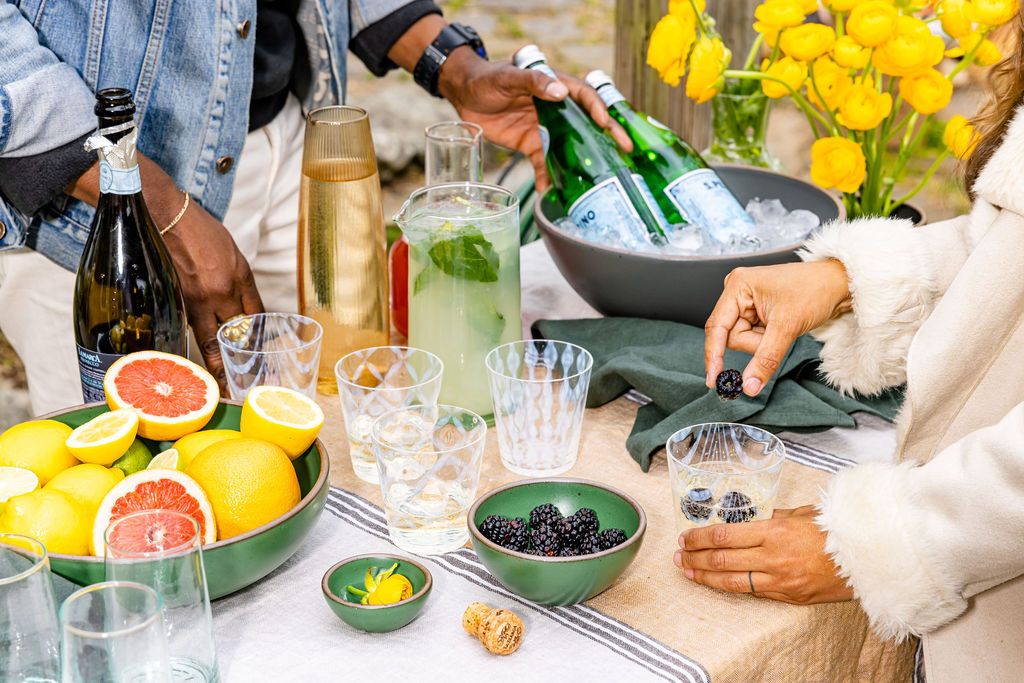 a table with assorted drinks and glasses as well as a bowl filled with ice and wine bottles and a separate bowl filled with whole and sliced citrus fruit