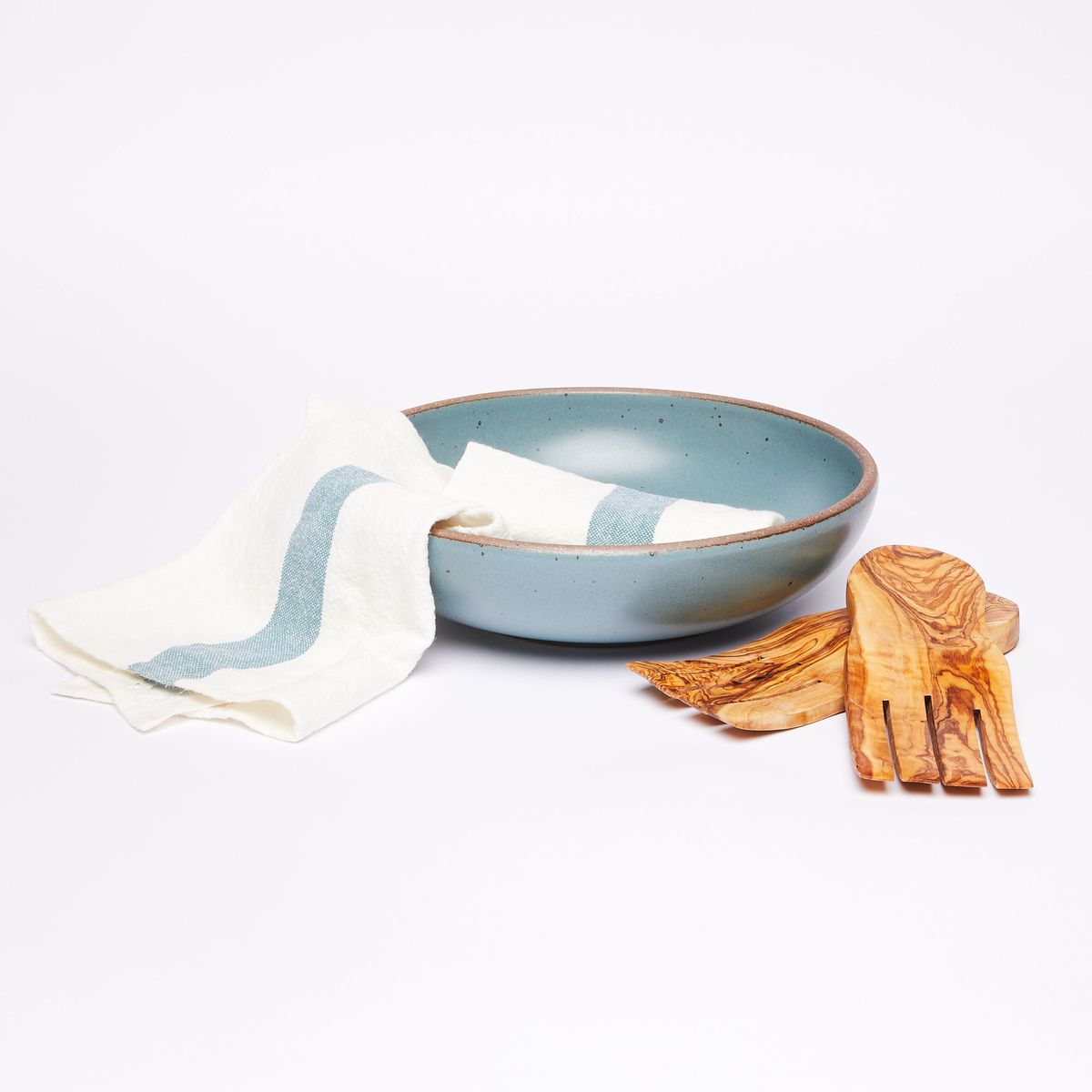 White tea towel with blue stripe on a large shallow turquoise serving bowl next to a pair of olivewood salad hands