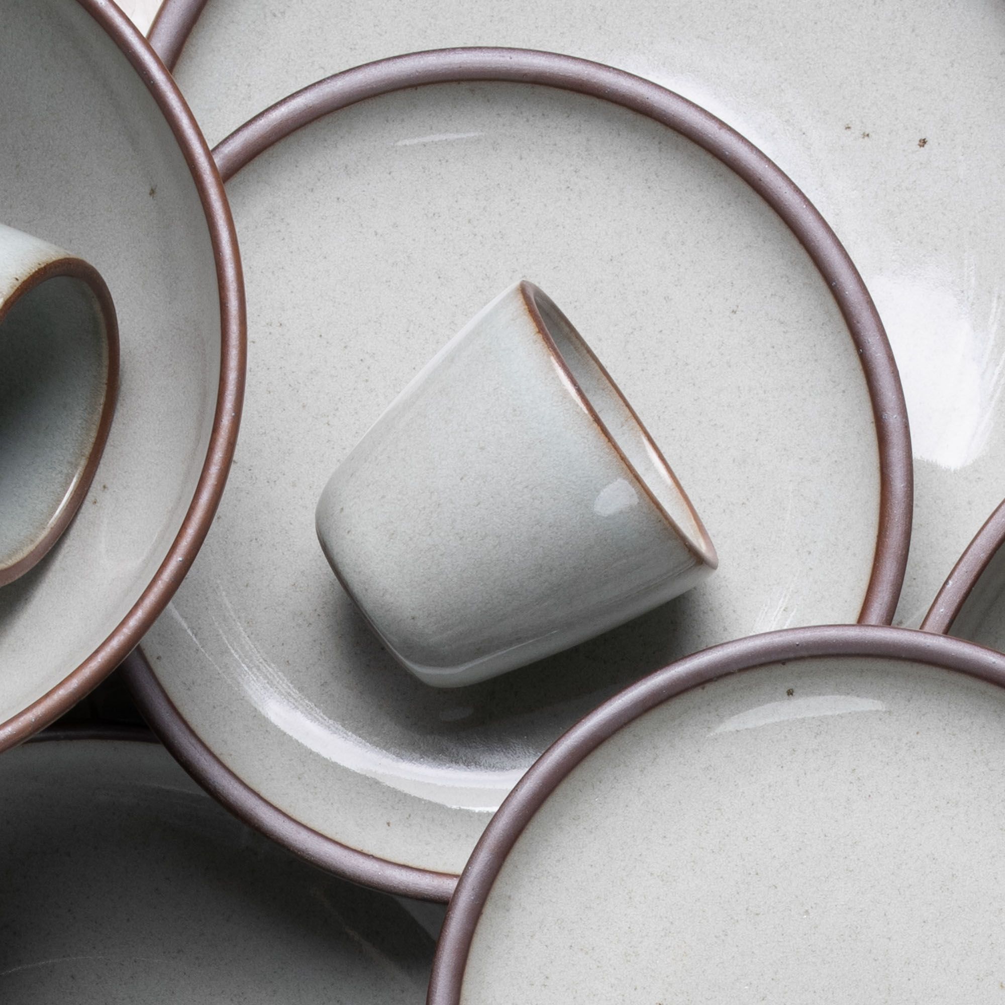 A short cup that tapers out to get wider at the top in a reflective icy off-white color featuring iron speckles, artfully arranged on other ceramic dinnerware forms.