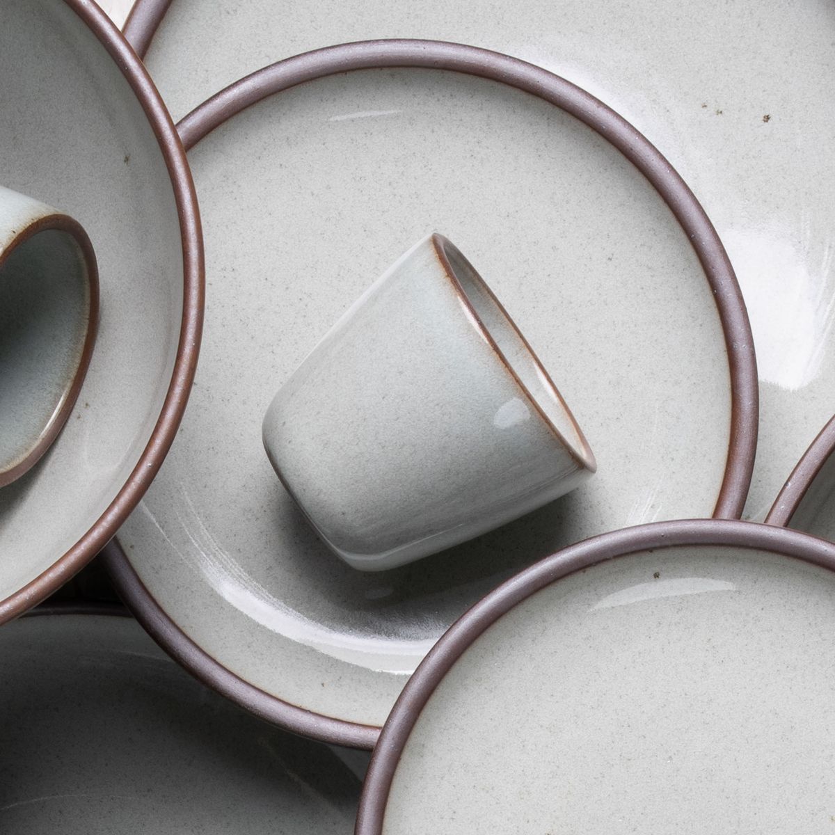 A short cup that tapers out to get wider at the top in a reflective icy off-white color featuring iron speckles, artfully arranged on other ceramic dinnerware forms.