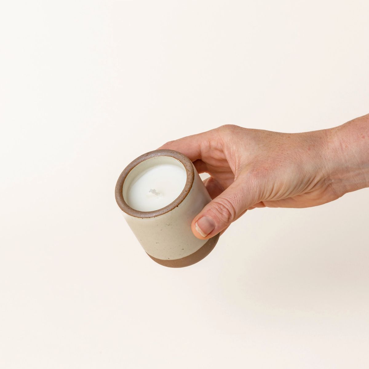 Hand holding a small ceramic vessel in a warm, tan-toned, off-white color with candle inside.