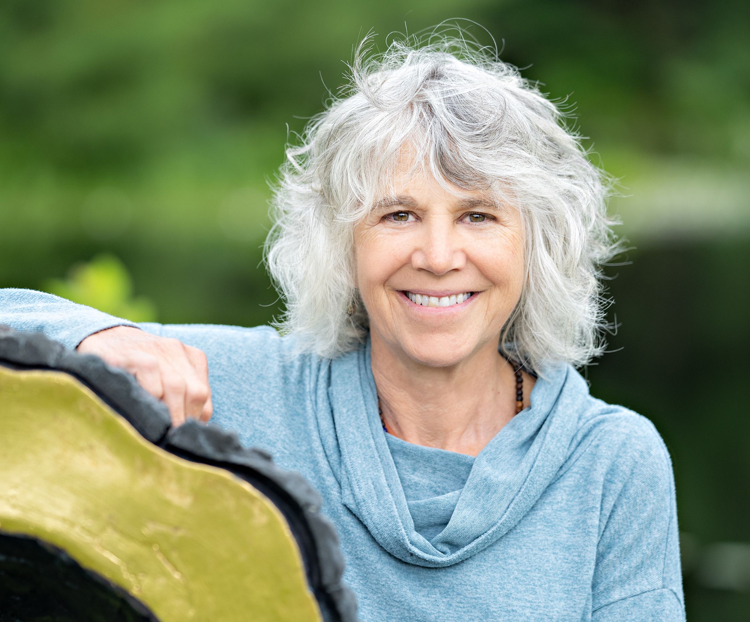 Linda Hoffman headshot, taken outside by a tree stump with green folliage in the background.
