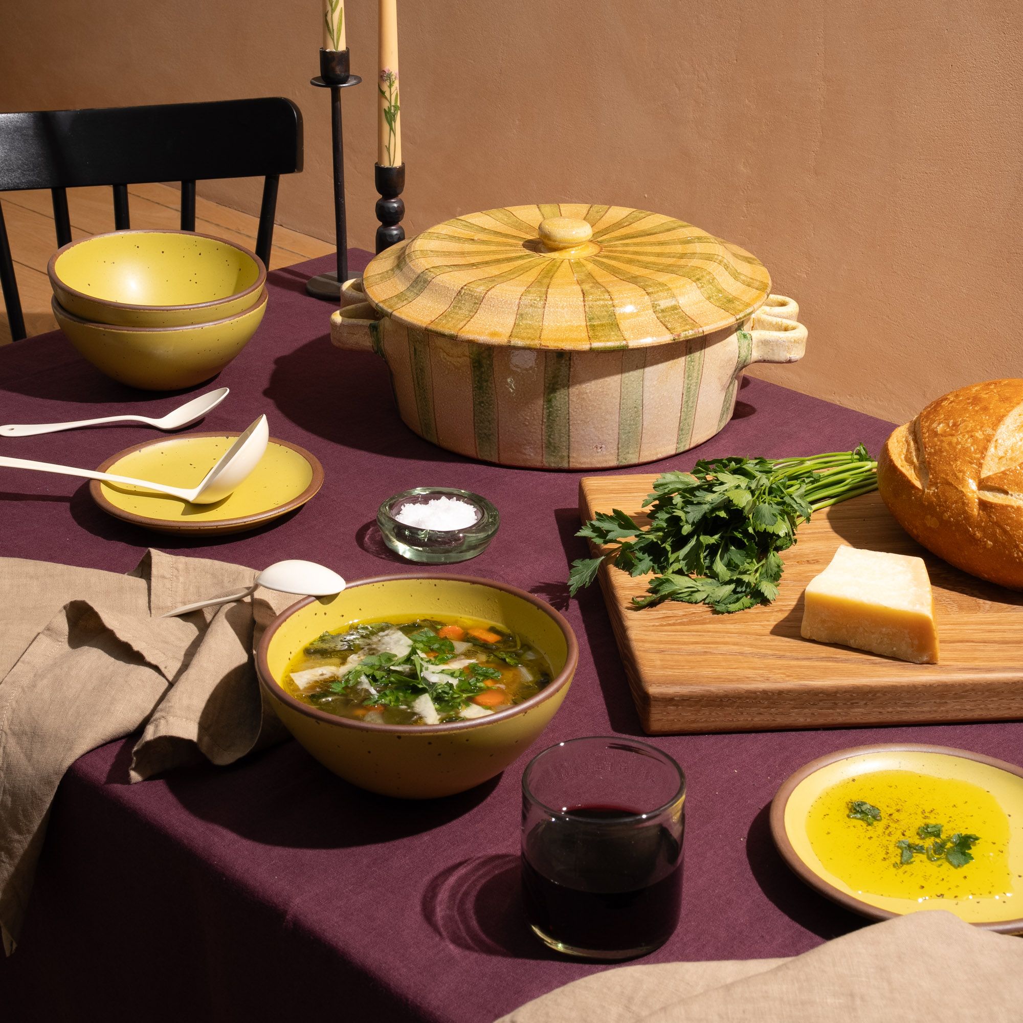 A table with with a large striped terracotta casserole dish, yellow soup bowls, a cutting board with bread, and more.