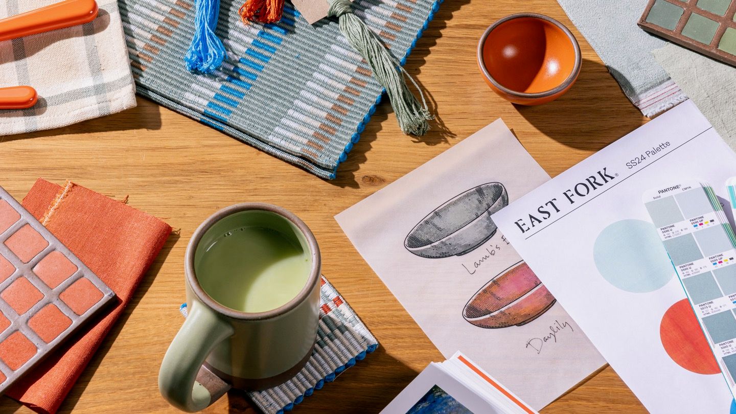 On a table, sits an artful display of textile samples, tile samples, ceramic mug and bowls, printouts and drawings -- all of a color story featuring sage green, bold orange, and a cerulean blue.