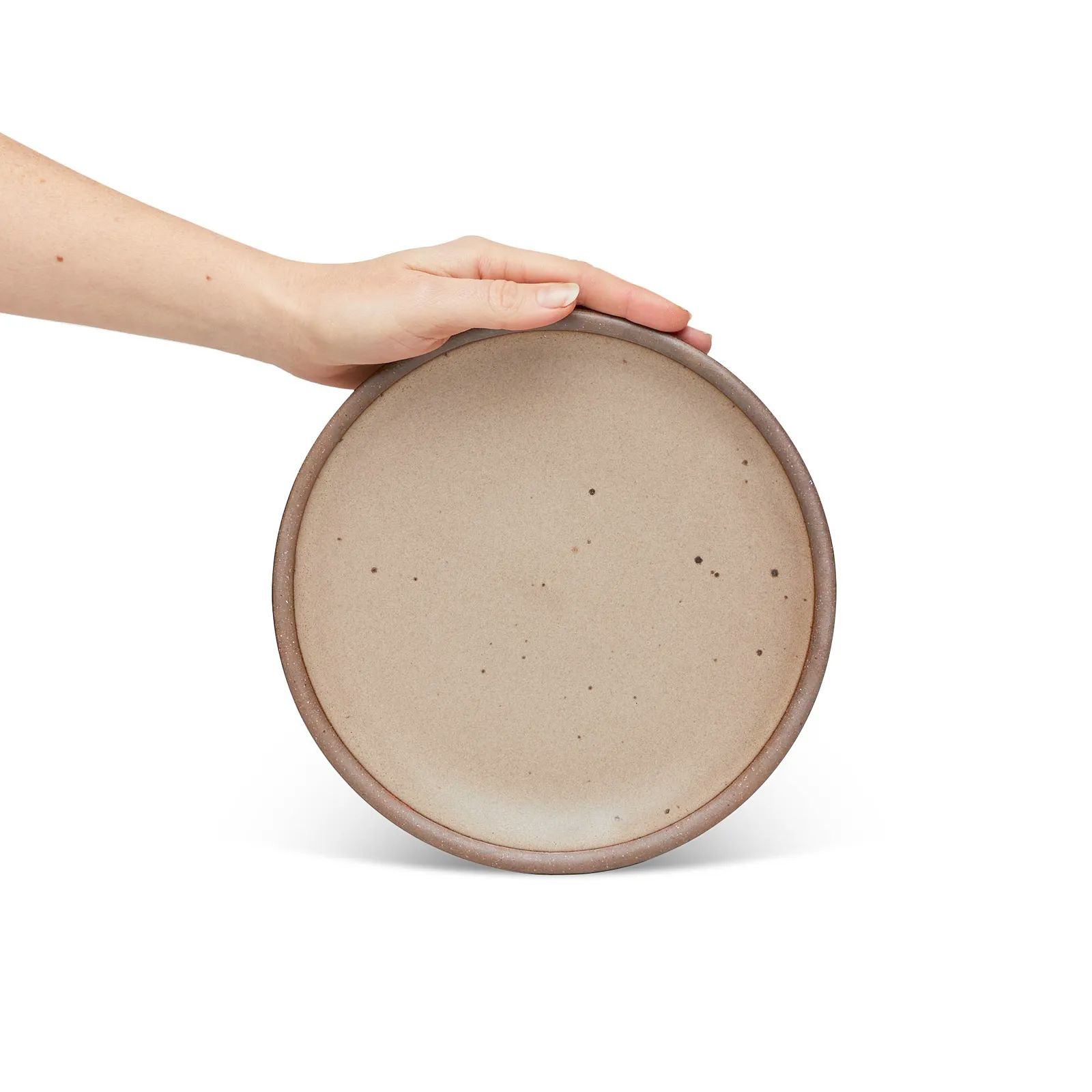 A hand holding a medium sized ceramic plate in a warm pale brown color featuring iron speckles and an unglazed rim.