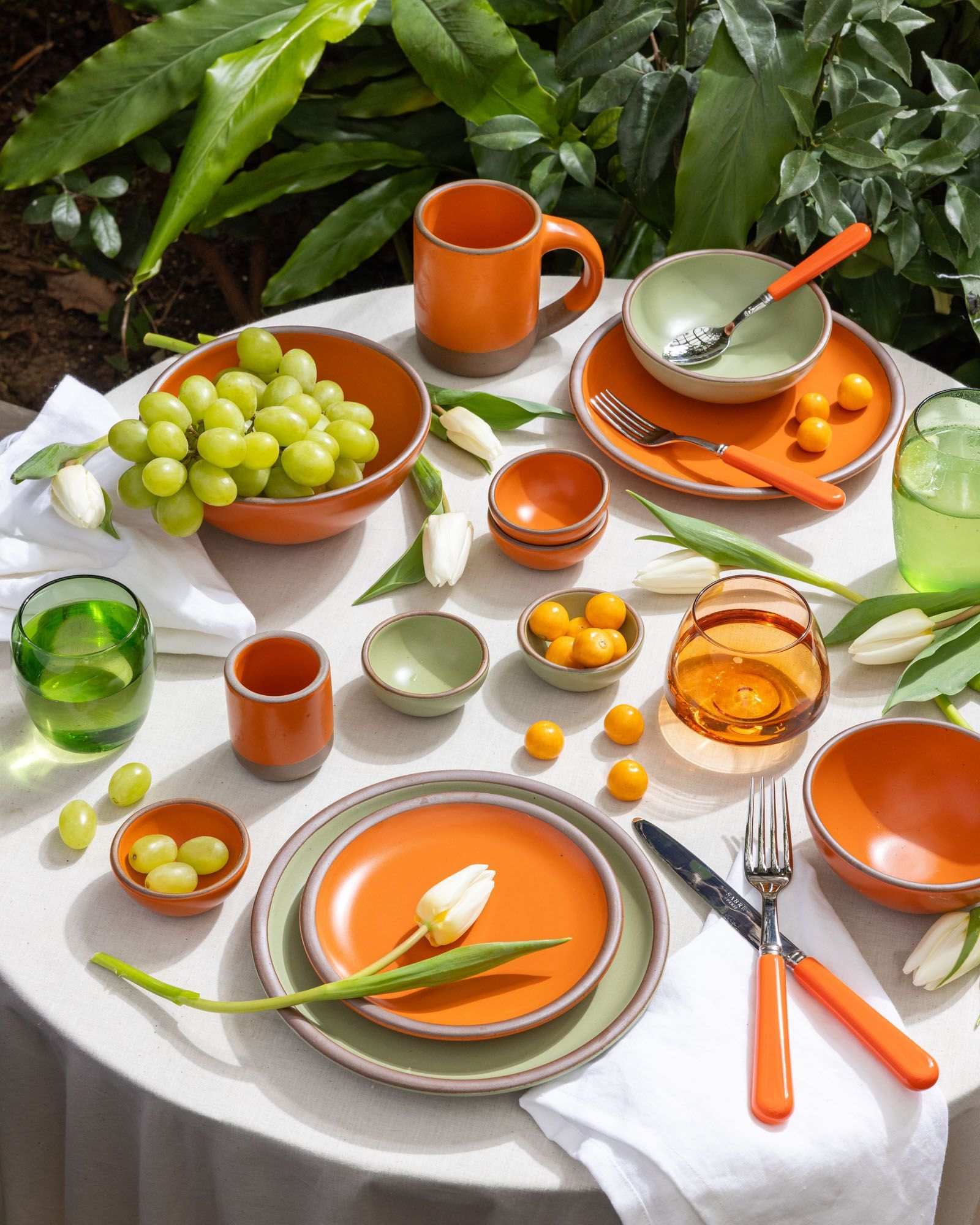 In an outdoor setting, a table is covered in ceramic dinnerware in a bold orange and calming sage green colors, surrounded by glasses, flatware and flowers in the same color palette.