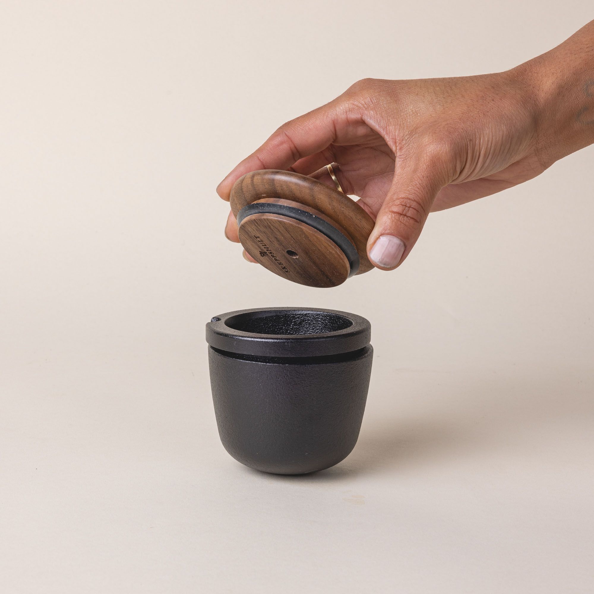 A short round cast iron jar with a wooden lid being held by a hand above it