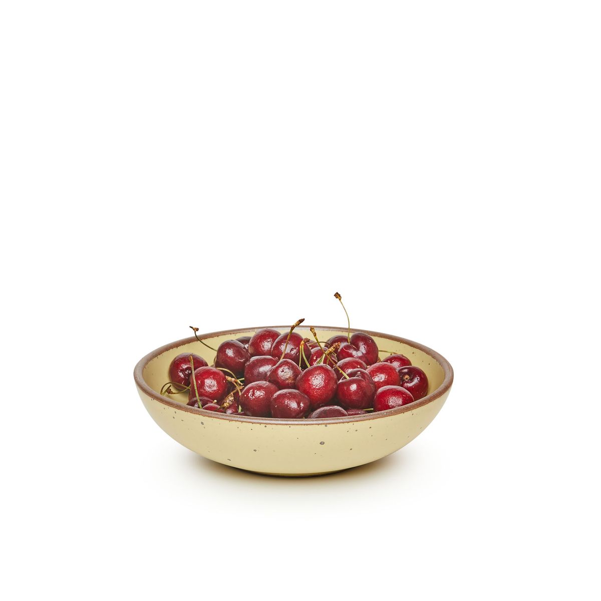 A dinner-sized shallow ceramic bowl in a light butter yellow color featuring iron speckles and an unglazed rim, filled with cherries