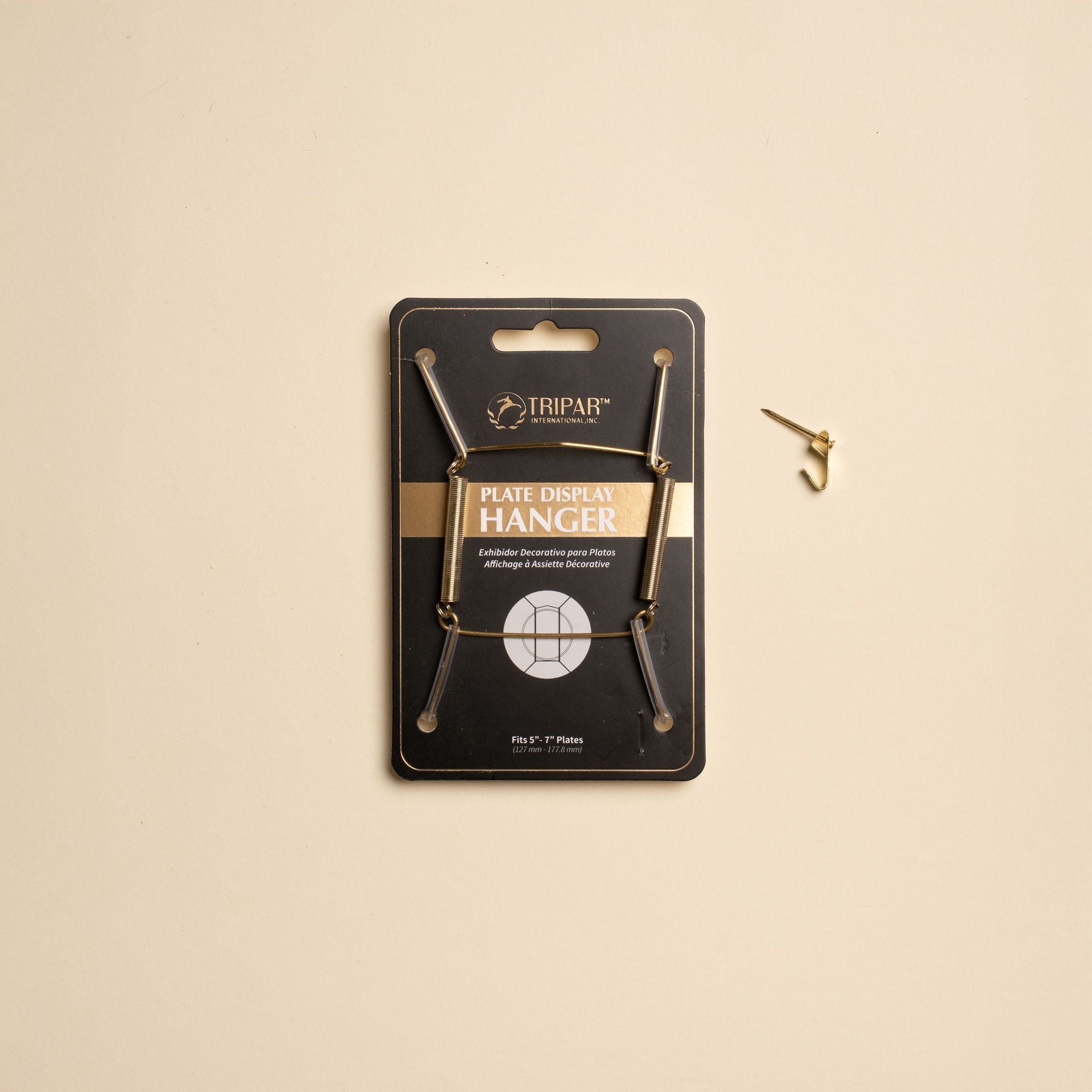 A brass plate hanger in its black cardboard package, with a nail off to the side.