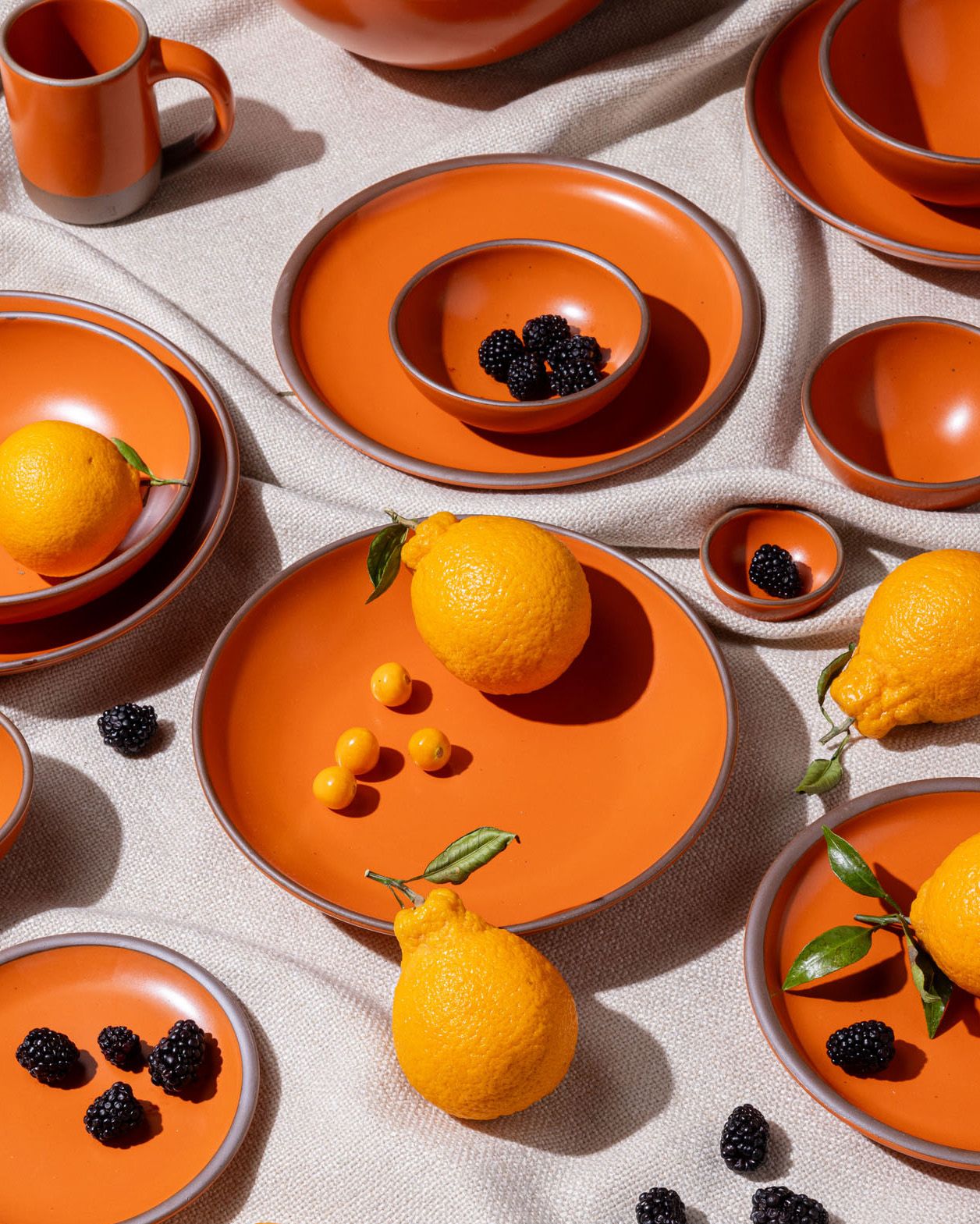 Ceramics plates and bowls of various sizes in a bold orange color, surrounded by blackberries, tomatoes, and oranges.