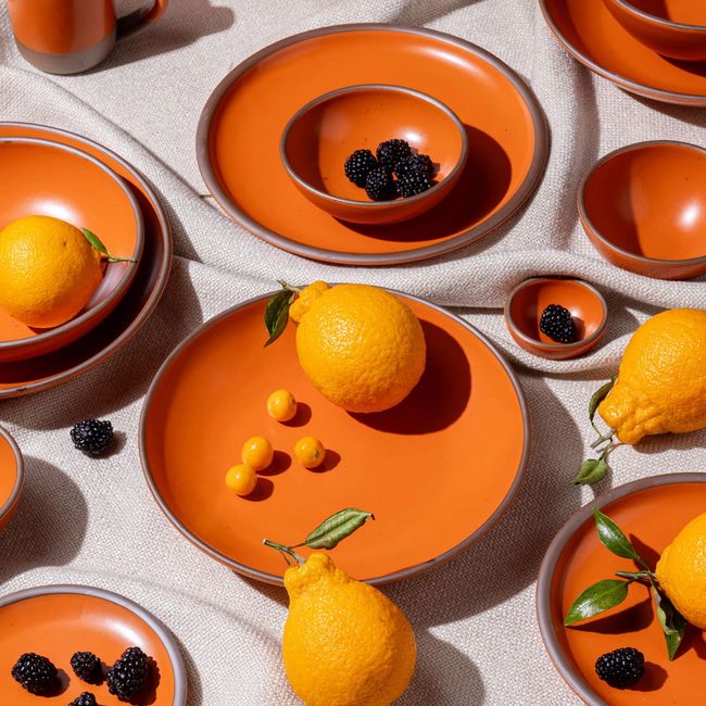 Ceramics plates and bowls of various sizes in a bold orange color, surrounded by blackberries, tomatoes, and oranges.